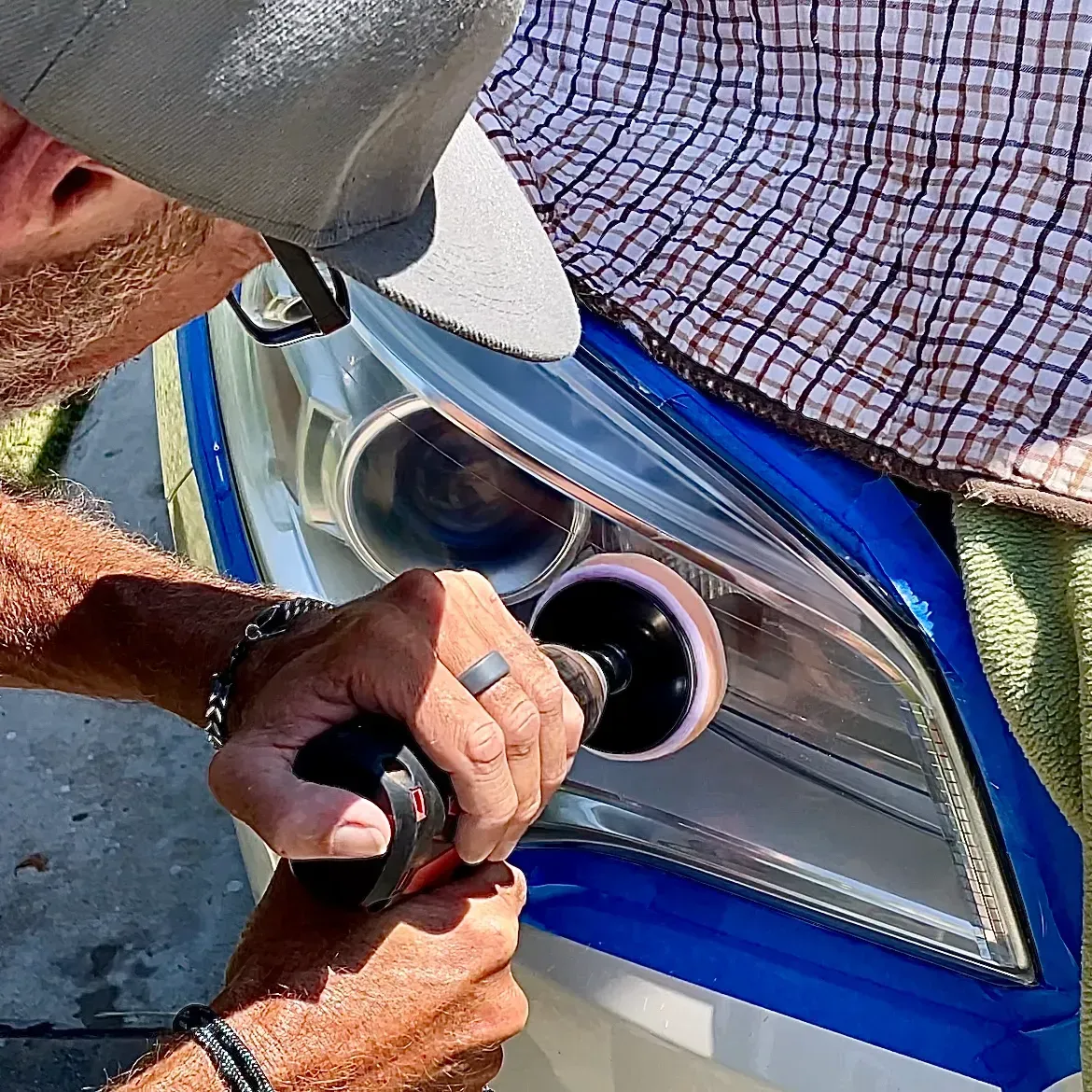 A person uses a power tool with a buffing pad to polish a car headlight that has been taped off with blue painter's tape.