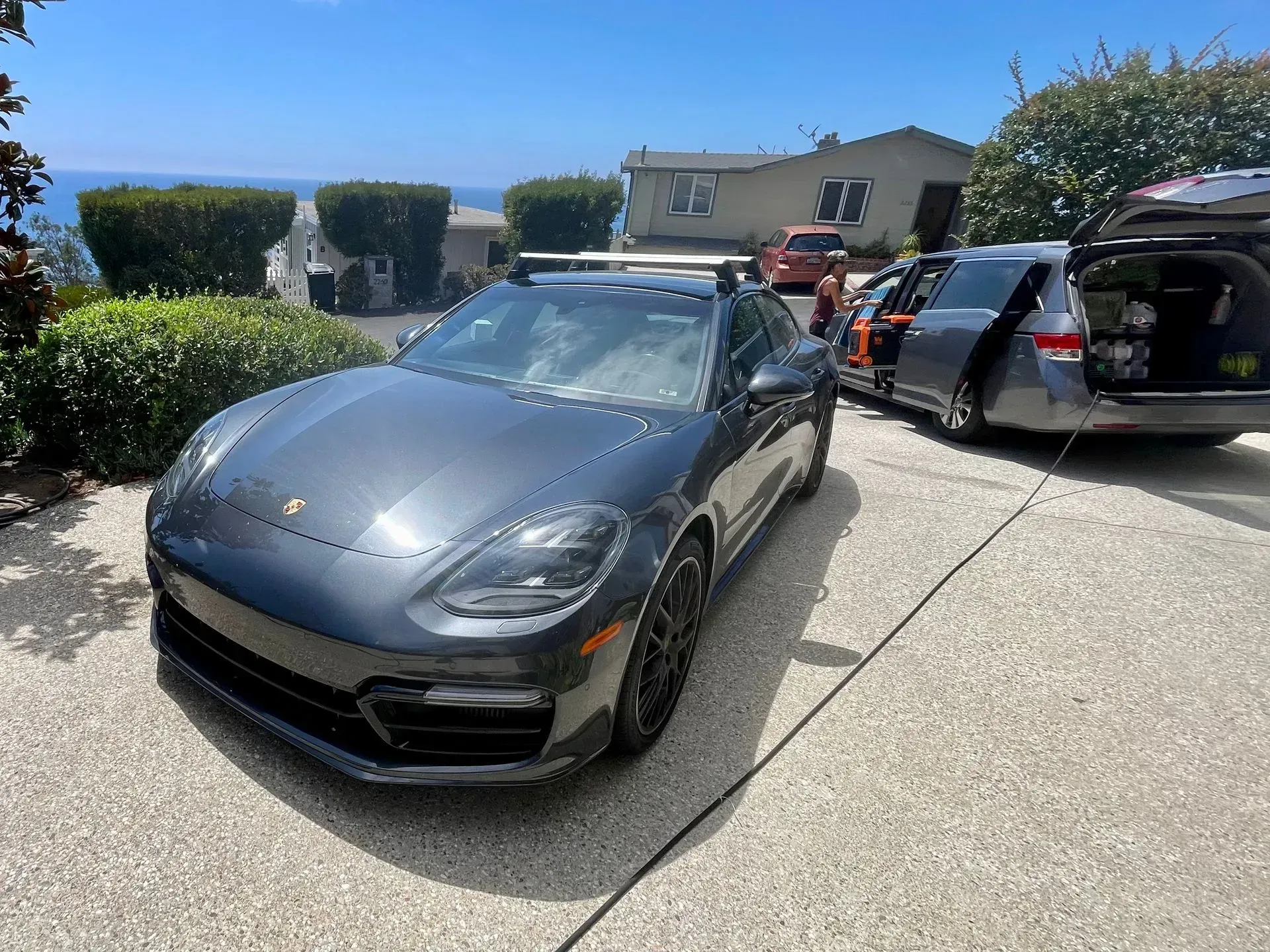 A dark grey Porsche parked on a driveway with a car parked behind it, rear hatch open, in a sunny residential setting.