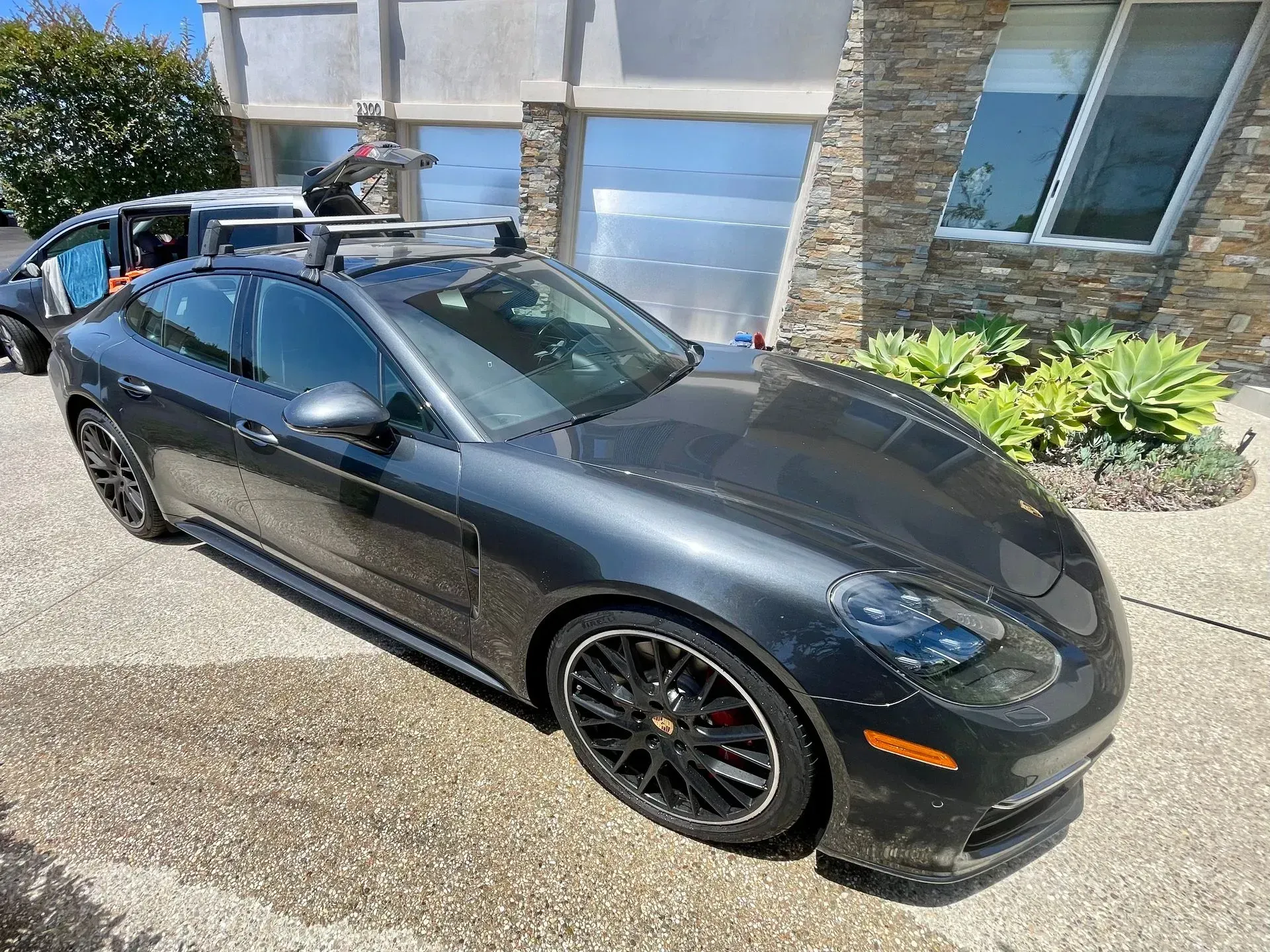 A dark gray Porsche sedan with a roof rack parked in a residential driveway on a sunny day.