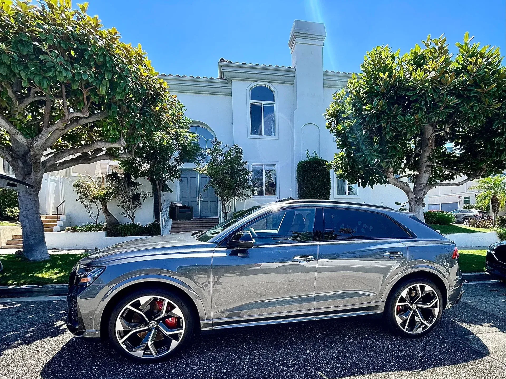 A grey Audi SUV parked on a gravel driveway in front of a white, multi-story stucco house with large trees.
