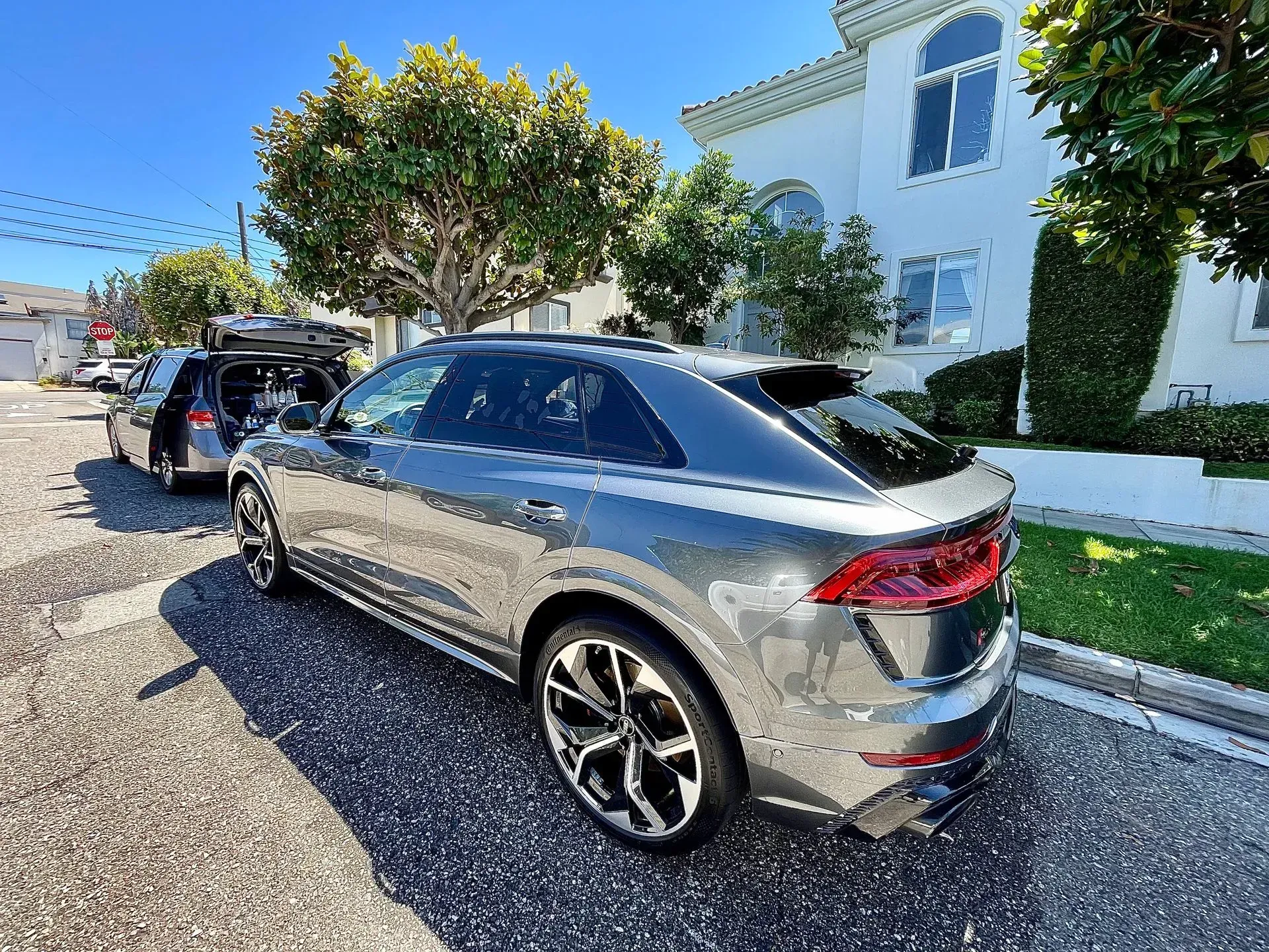 A grey Audi SUV parked on a sunny residential street in front of a white house, with another car visible behind it.