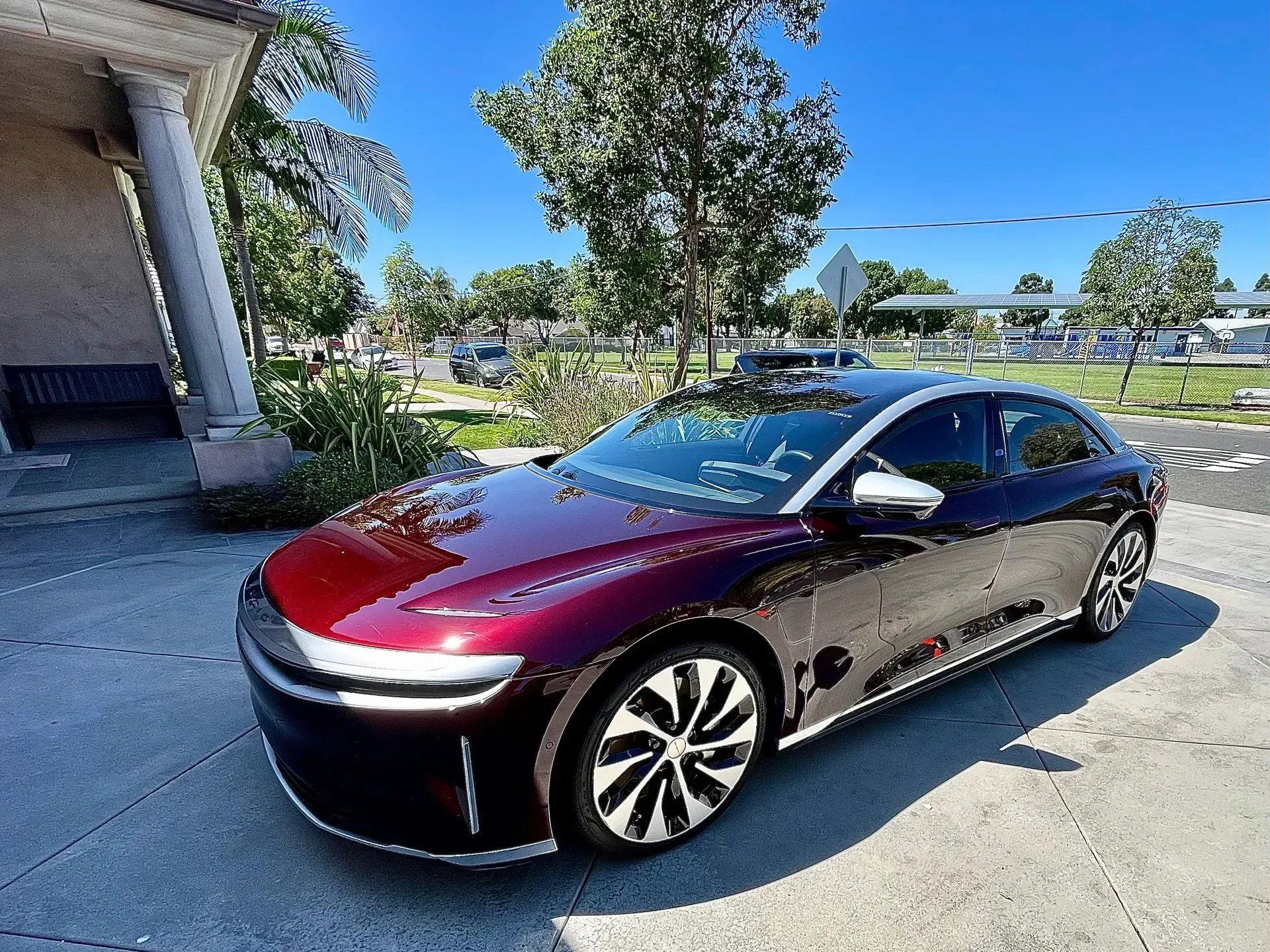 A dark metallic red Lucid Air electric sedan parked on a concrete lot near a building pillar under a clear blue sky.
