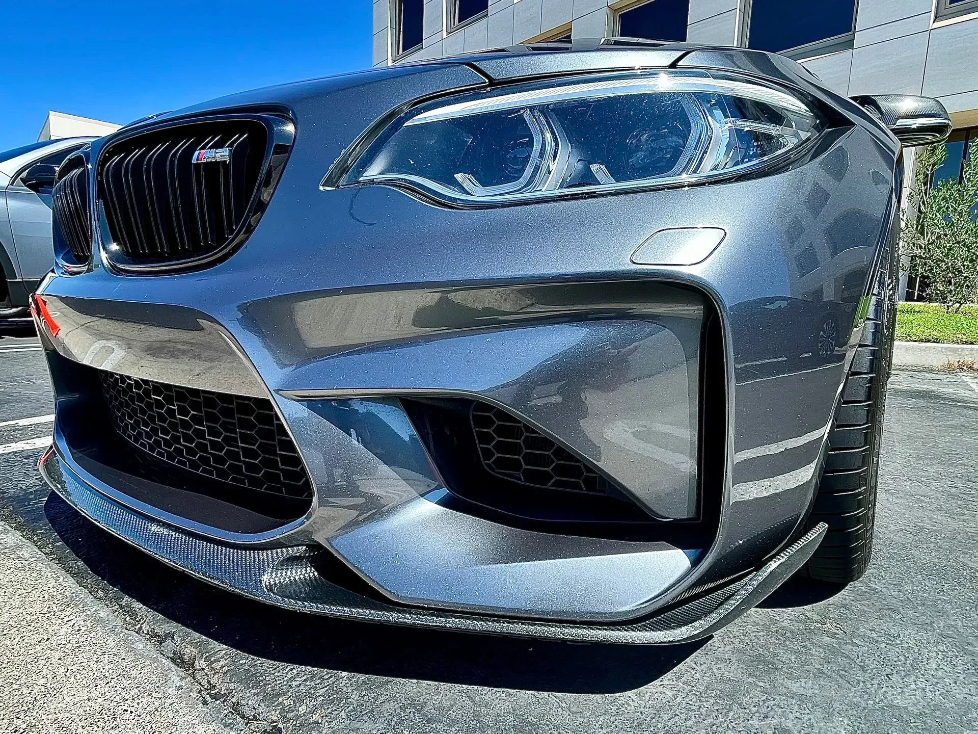 A close-up, low-angle shot of the front bumper and headlight of a metallic grey BMW M2 sports car parked outdoors.