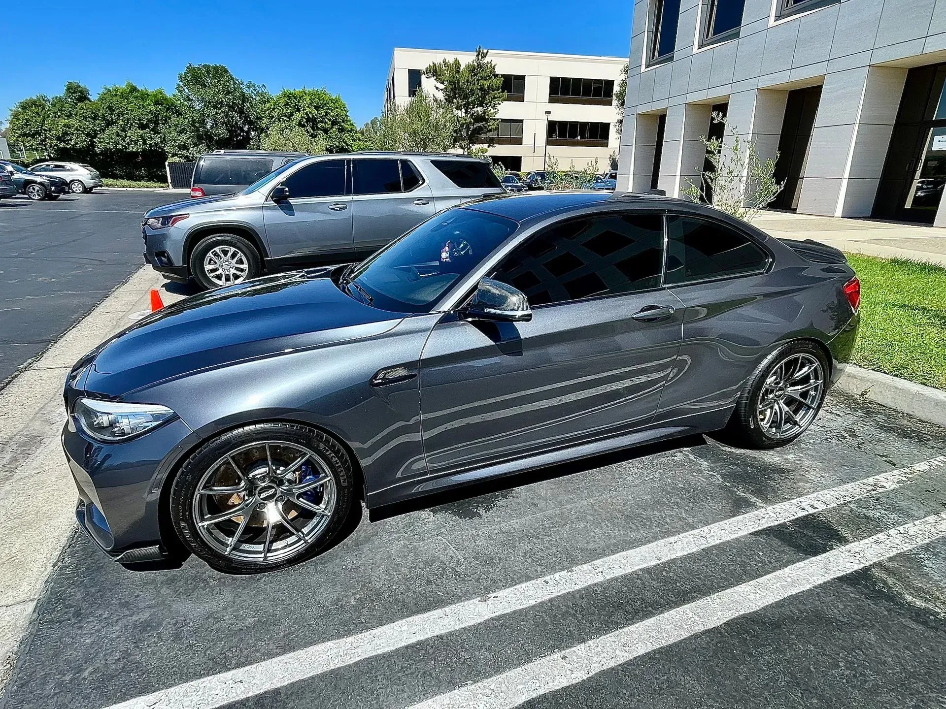 A dark grey BMW M2 coupe parked in a paved lot beside a light-colored building under a clear blue sky.