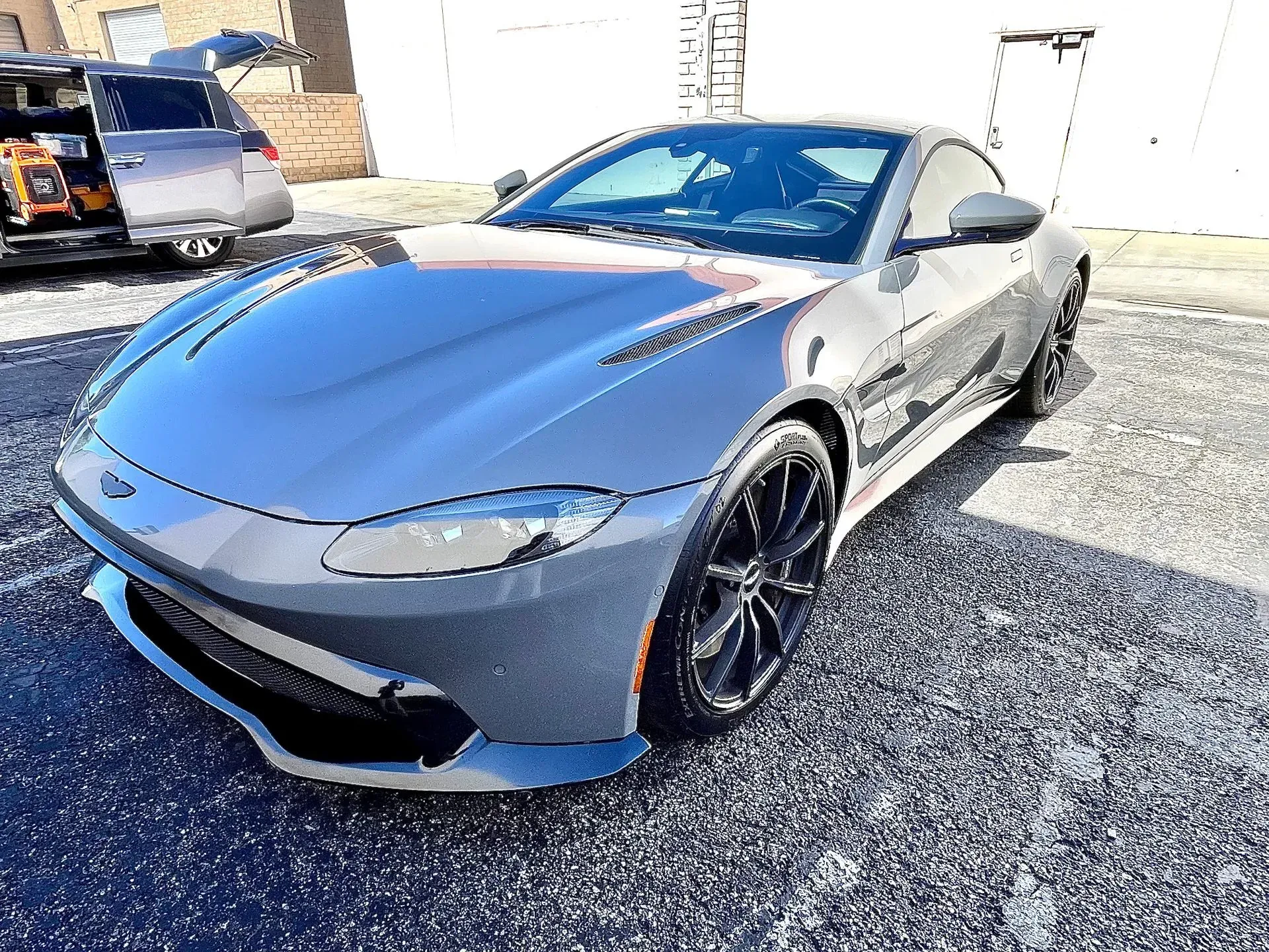 A grey Aston Martin sports car parked on an asphalt lot with a van visible in the background.