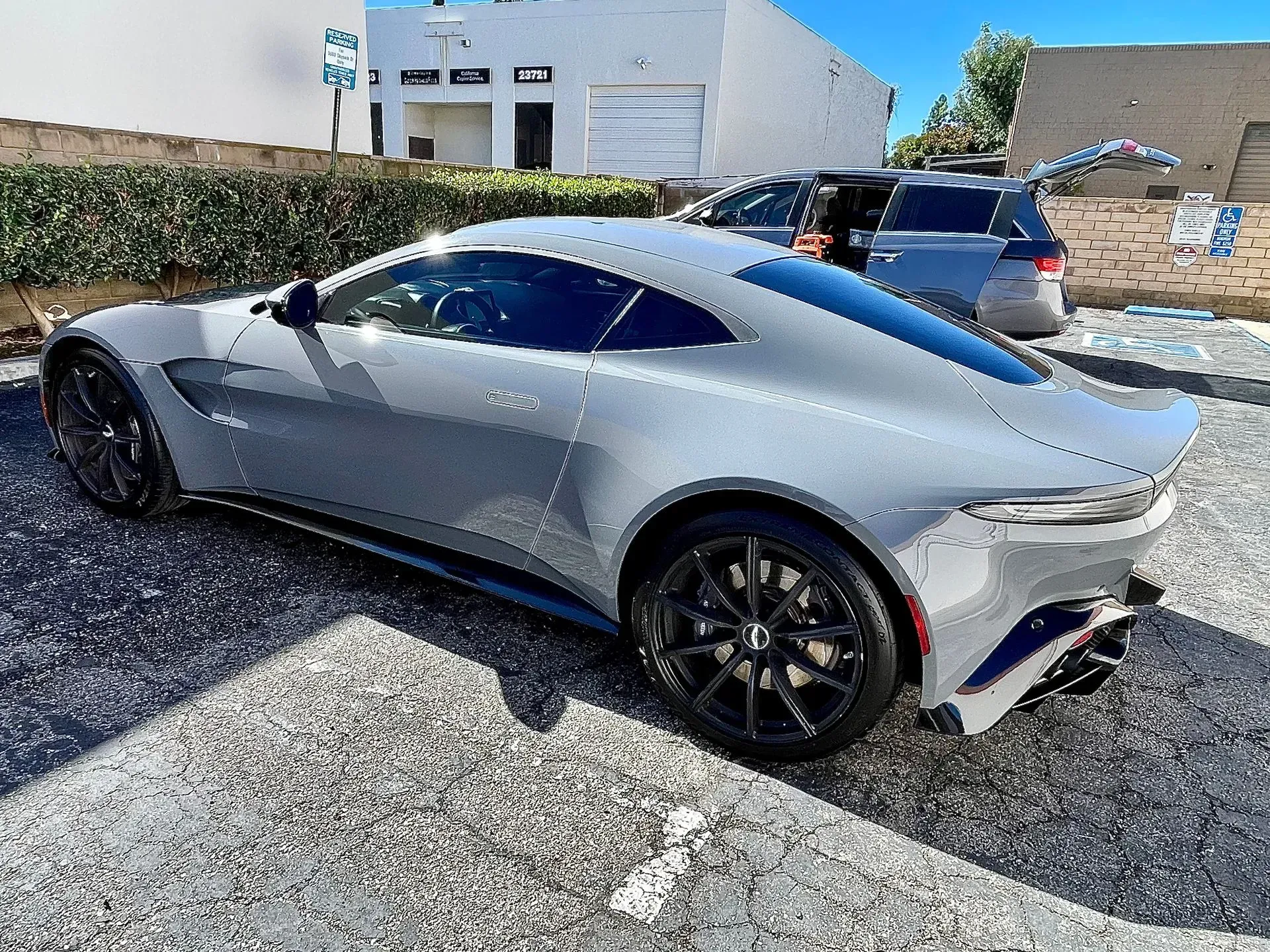 A sleek silver Aston Martin coupe parked on a gravel lot during a sunny day.