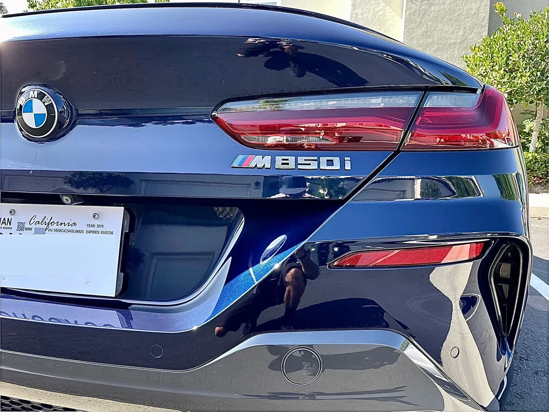 Rear view of a dark blue BMW M850i showing the logo, taillight, and license plate area in an outdoor setting.