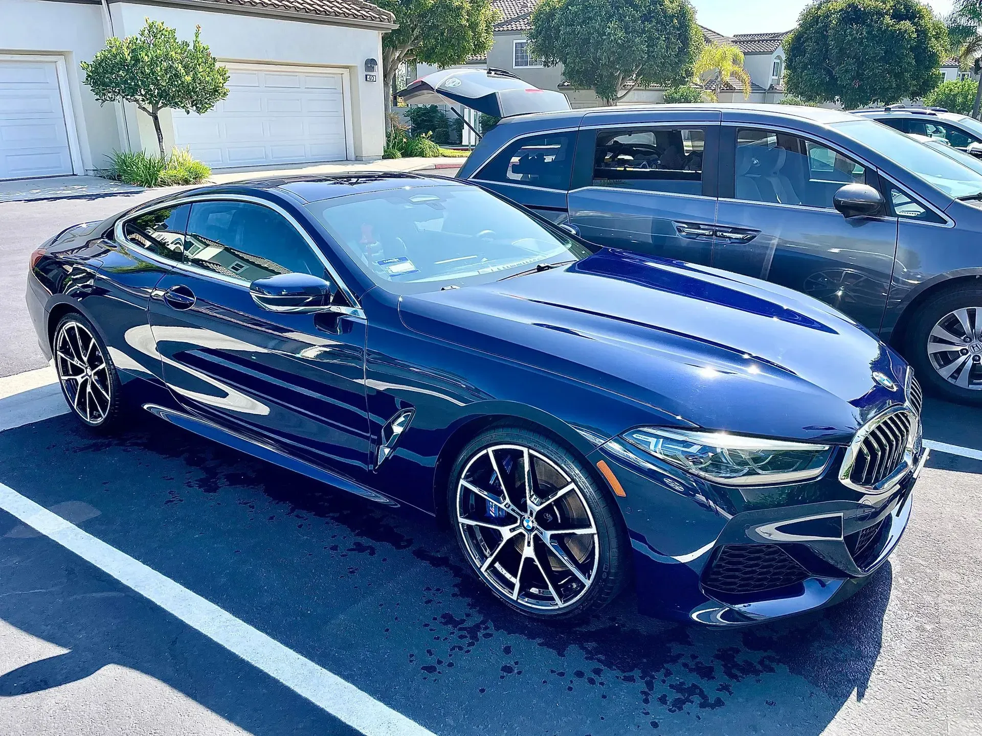 A dark blue BMW 8 Series coupe parked in an outdoor lot on a sunny day next to a grey minivan.