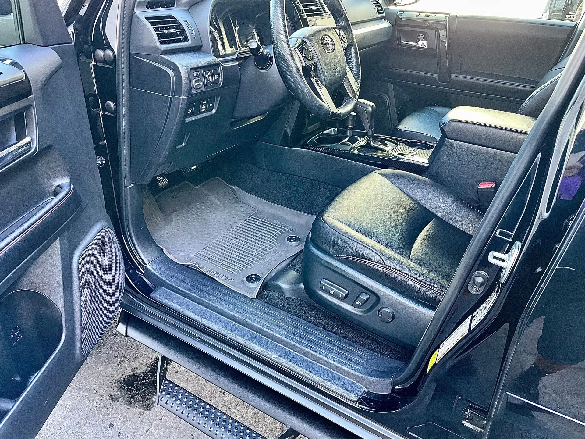 The interior of a black vehicle showing the driver-side door open to a leather seat, steering wheel, and rubber floor mat.