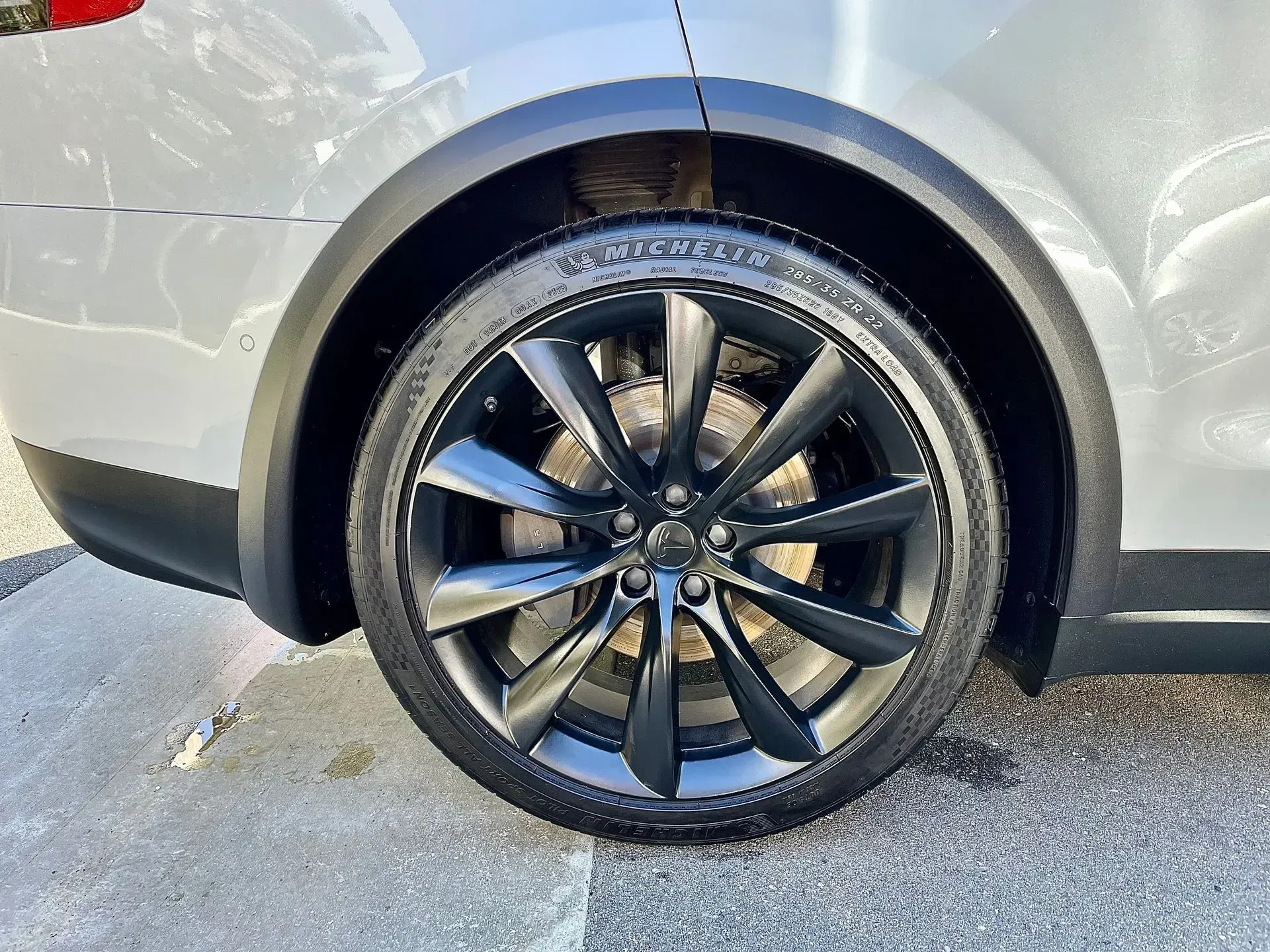 A close-up view of a matte black car wheel with thin spokes on a grey vehicle parked on asphalt.