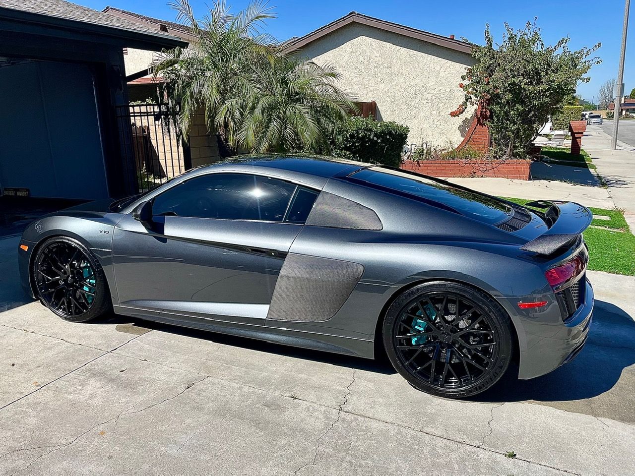 a gray supercar Audi R8 with carbon fiber body parts, glossy black wheels and bright teal brake calipers, sits gleaming in the sun parked in a residential driveway, freshly detailed