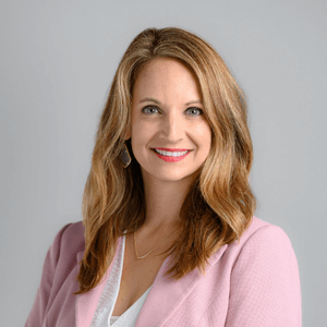 Woman with long blonde hair smiles, wearing a pink blazer and white shirt against a gray backdrop.