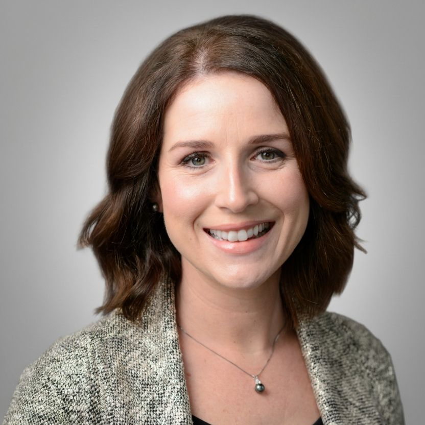 Woman with brown hair, smiling, wearing a patterned blazer and necklace.