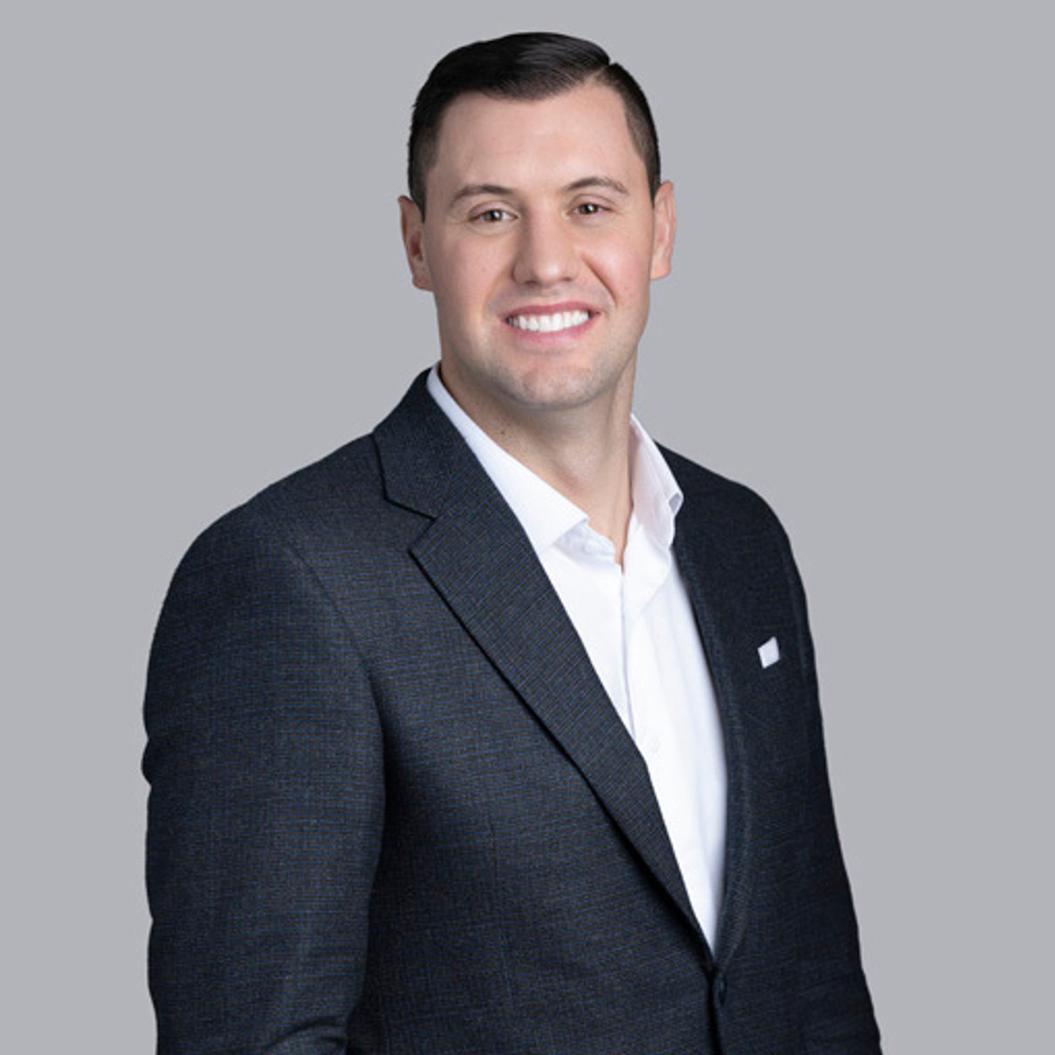 Man in dark blazer and white shirt smiles at the camera against a gray background.