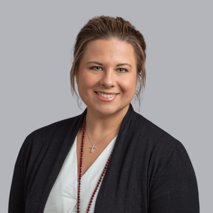 Woman with brown hair smiles, wearing a black cardigan, white top, and beaded necklace. Gray background.