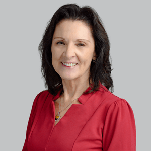 Woman with long blonde hair smiles, wearing a pink blazer and white shirt against a gray backdrop.