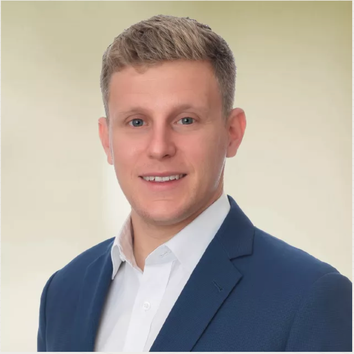 Man in a navy blazer and white shirt smiles, light hair, against a soft yellow background.