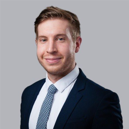 Man in a navy suit and tie, smiling, against a gray background.