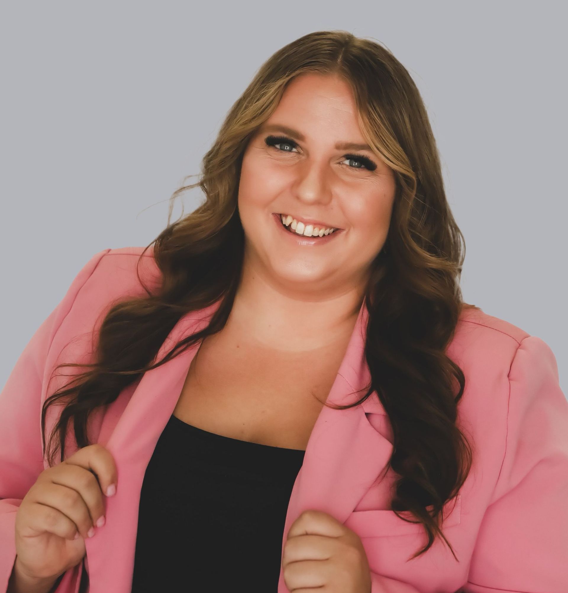 Woman in a pink blazer smiles broadly, hands on lapels, against a gray background.