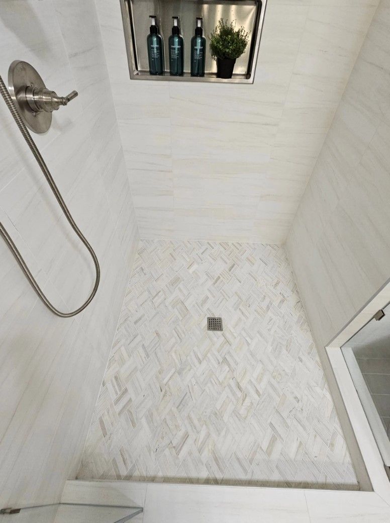 White tiled shower with herringbone floor, built-in shelf with bottles, and a handheld showerhead.