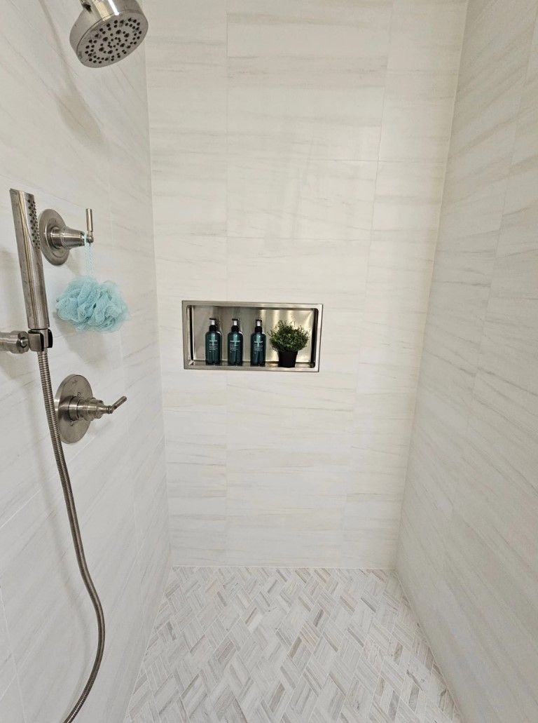 A white-tiled shower with a built-in shelf holding toiletries. Herringbone floor design.