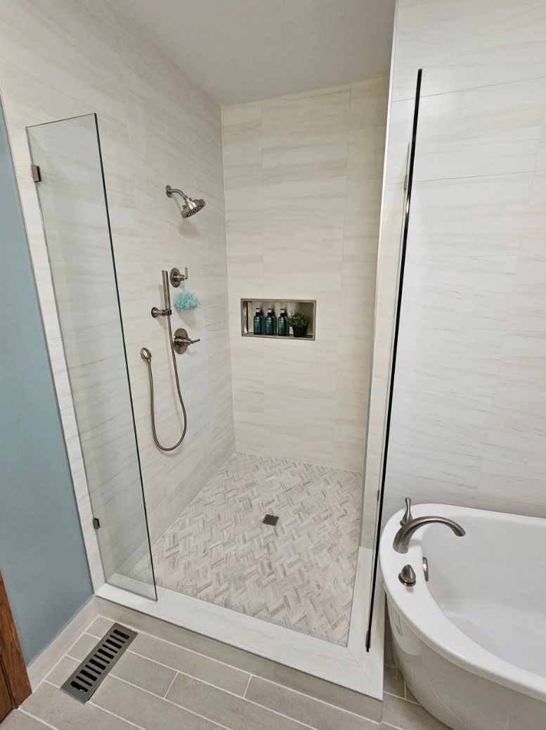 Shower with glass door and tiled walls, next to a bathtub. Light gray and white tones.