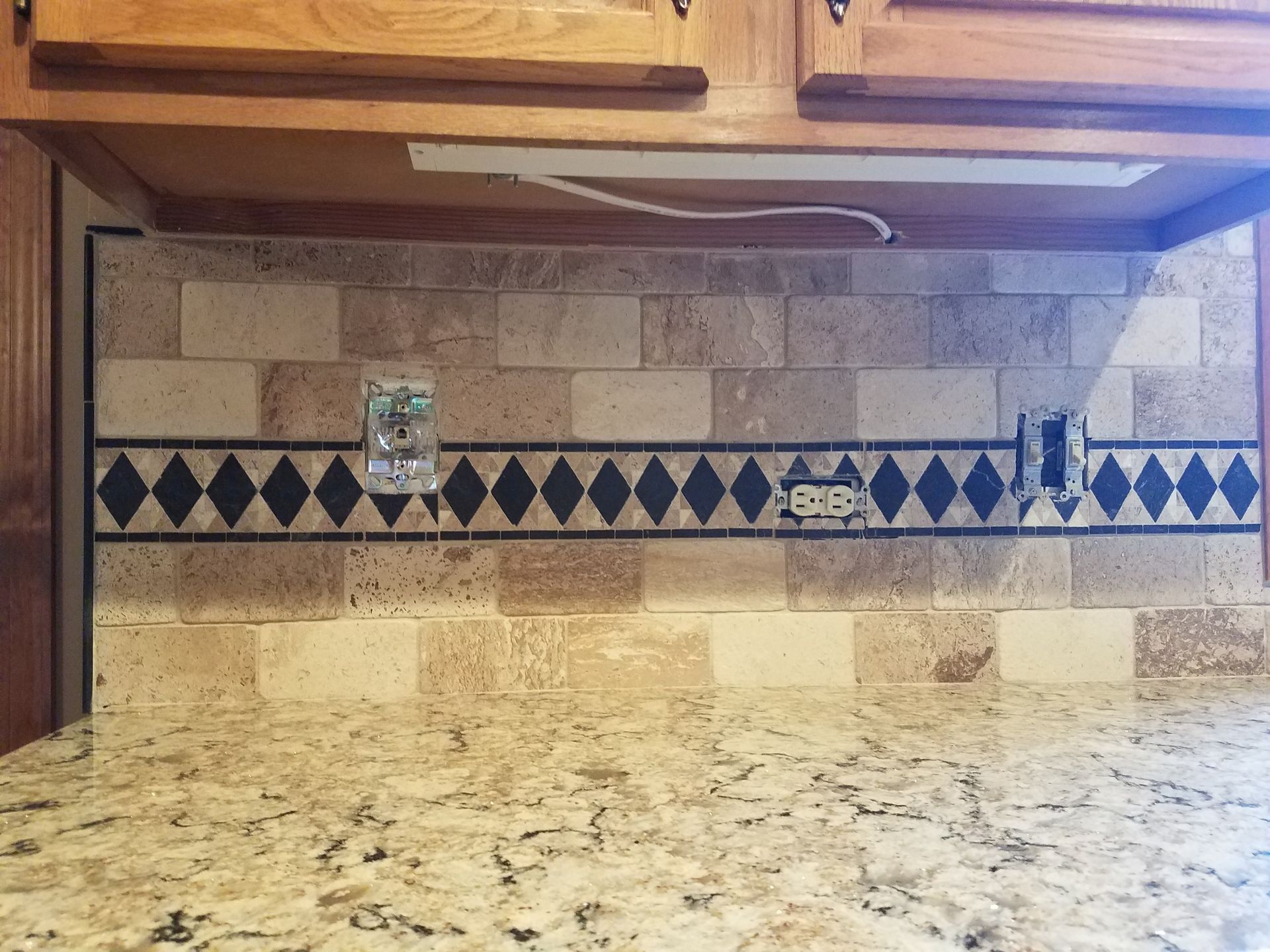 Kitchen backsplash with granite countertop, beige tile, a decorative black and white diamond border, and outlets.