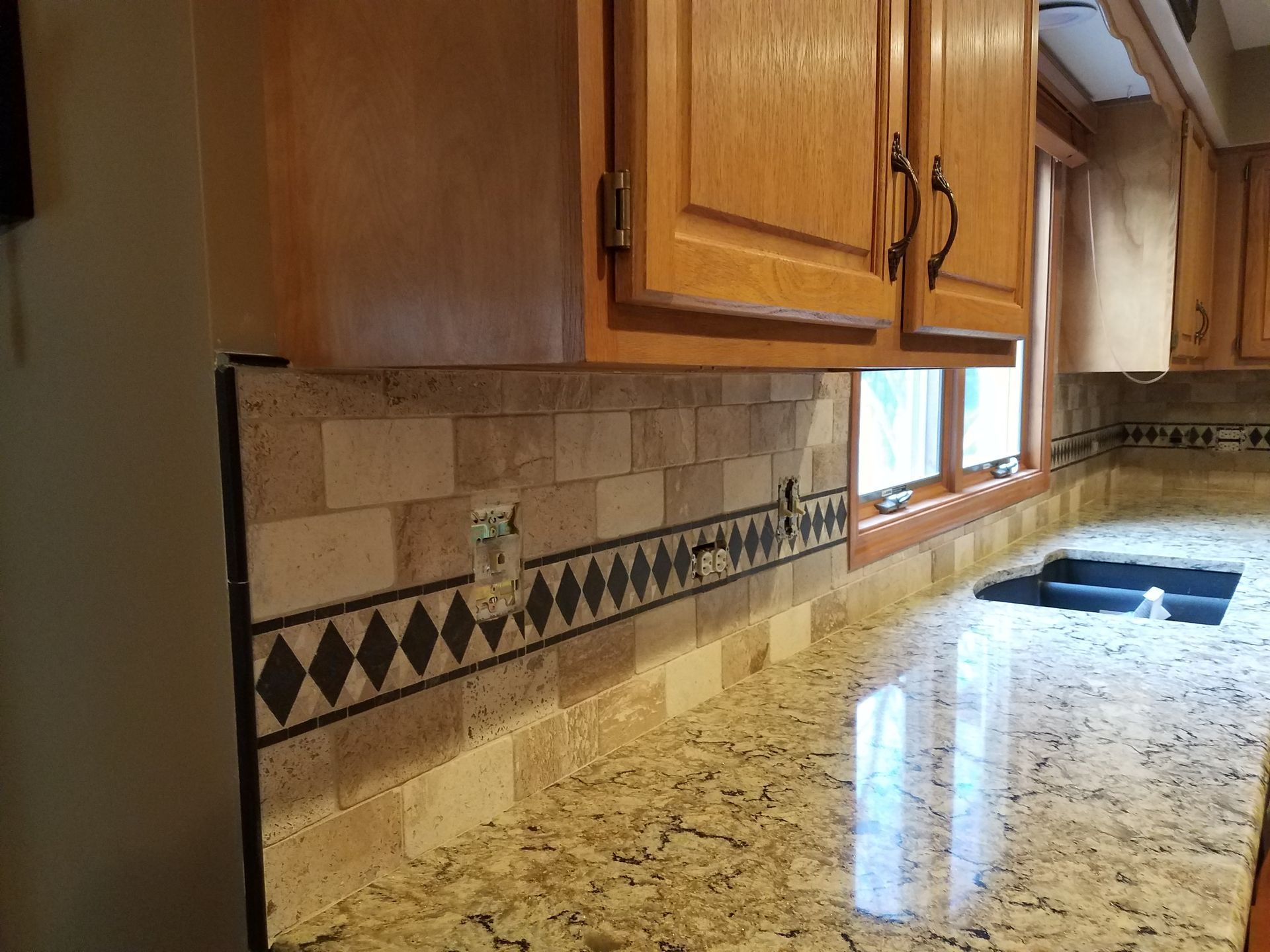 Kitchen backsplash with tan tile, black diamond border, granite countertop, and wooden cabinets.