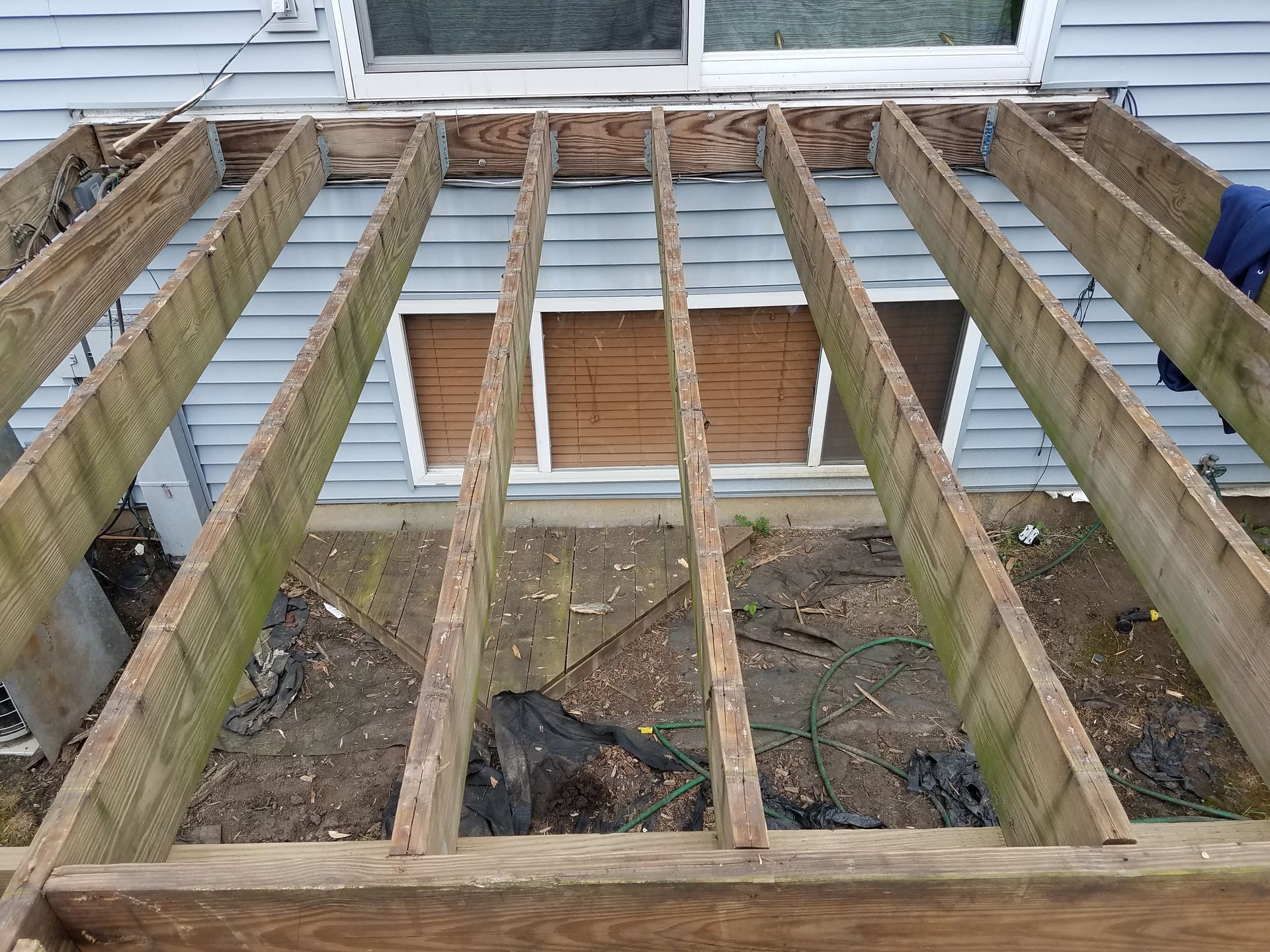 Overhead view of a wooden deck frame, missing boards. The weathered wood is in disrepair near a blue-sided house.