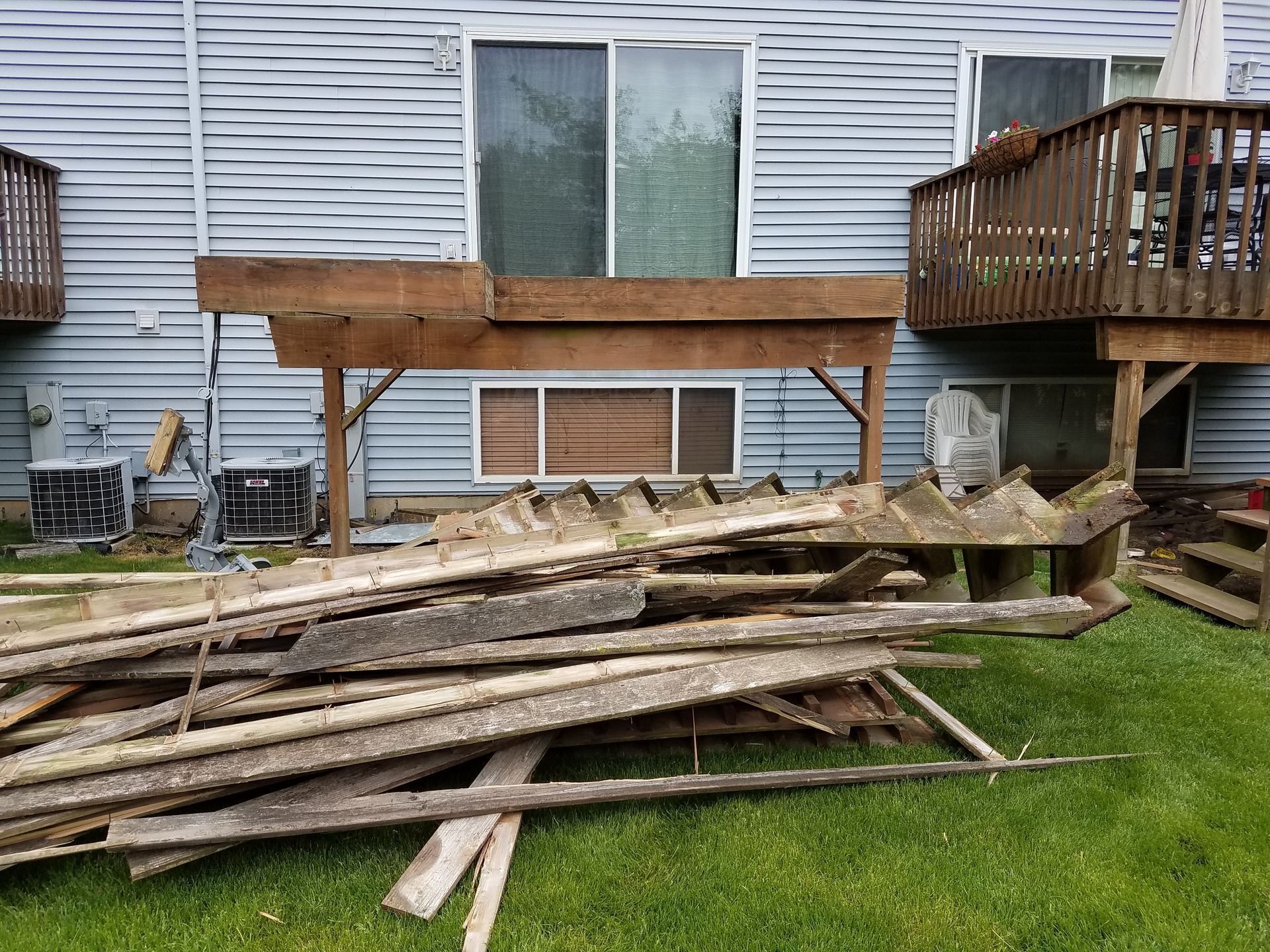 Pile of weathered wooden planks in front of a partially-built deck attached to a blue-sided building.