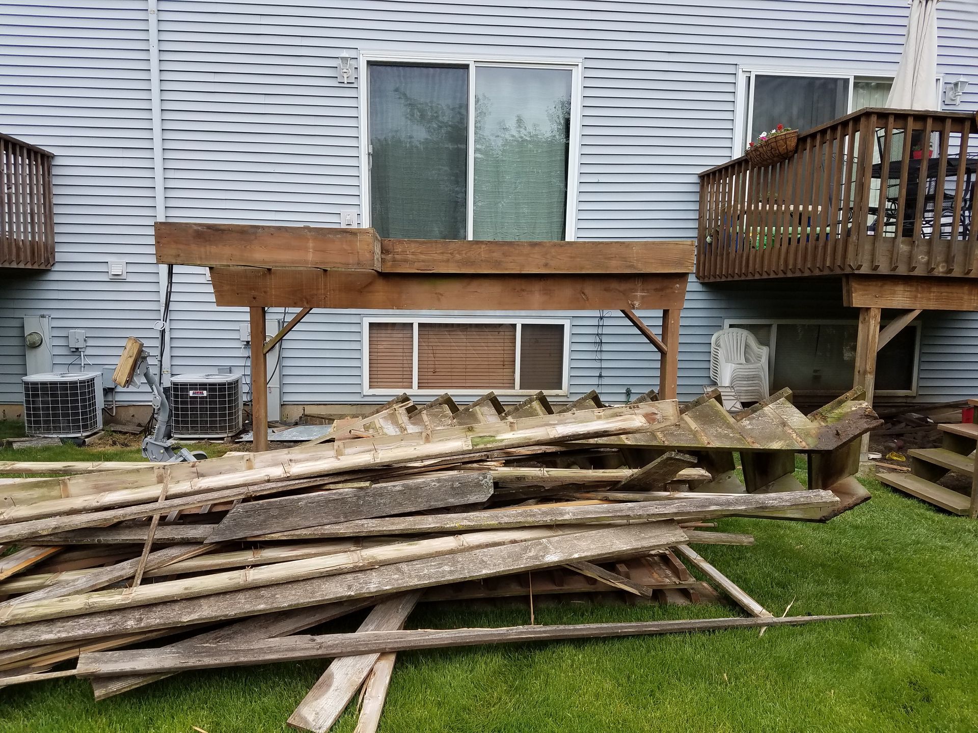Pile of weathered wood in front of a house. Partially dismantled deck with support beams visible.