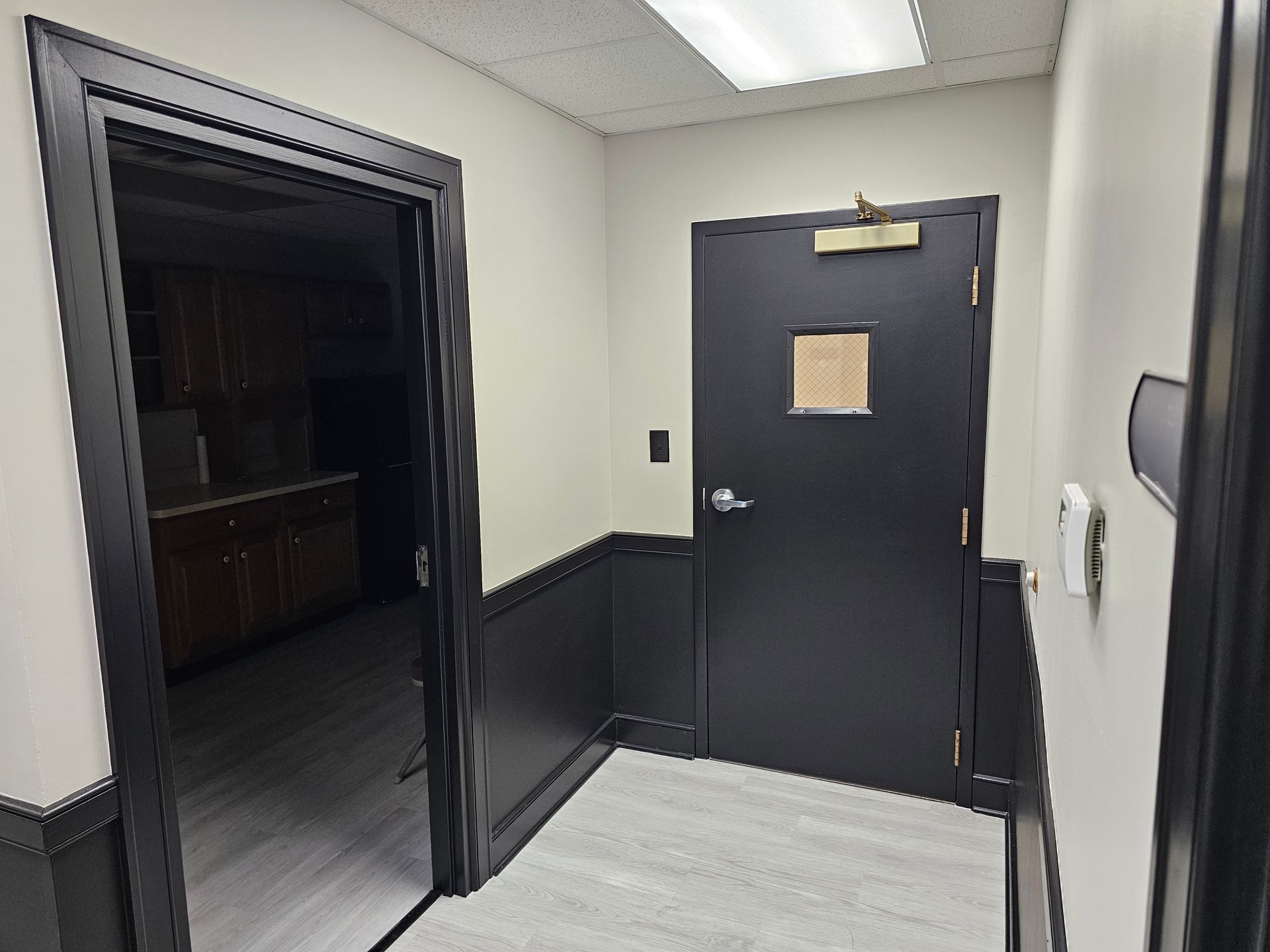 Hallway with black door, door frame, and wainscoting against white and tan walls. Open door reveals room with cabinetry.