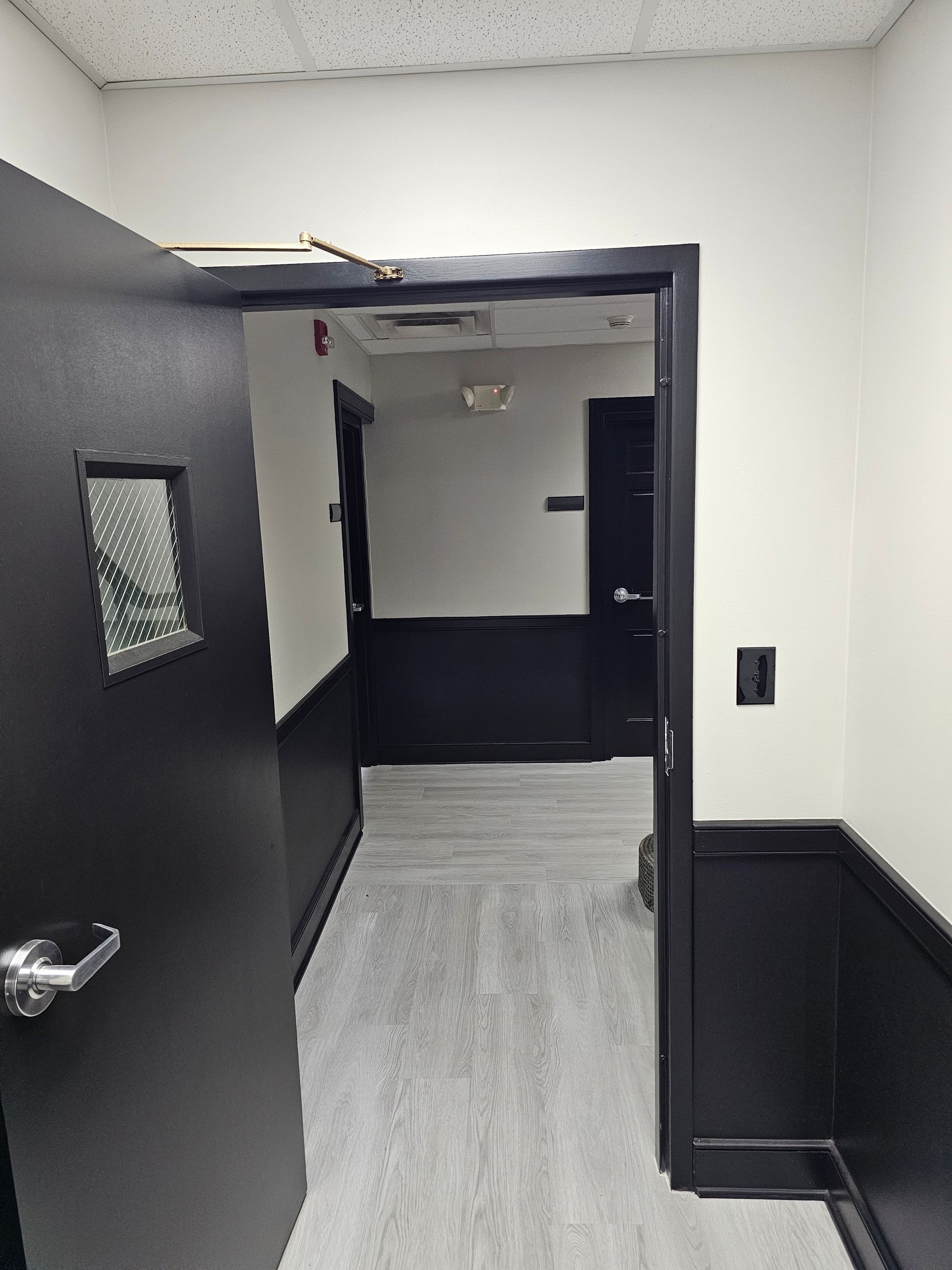 Open black door, leading into a hallway with black wainscoting, white walls, and light gray floor.