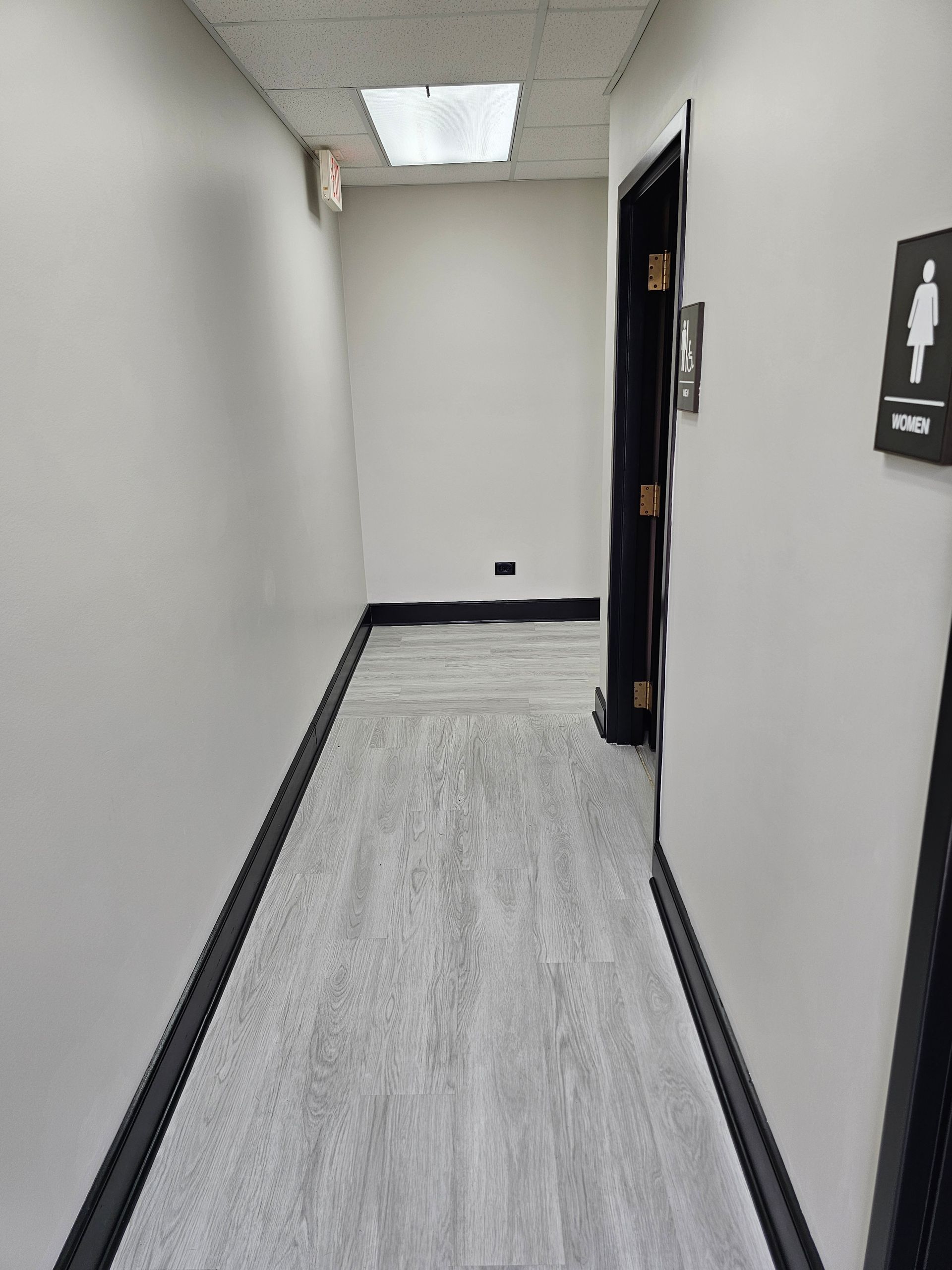Narrow hallway with light gray flooring, white walls, and a dark door. A restroom sign is visible.