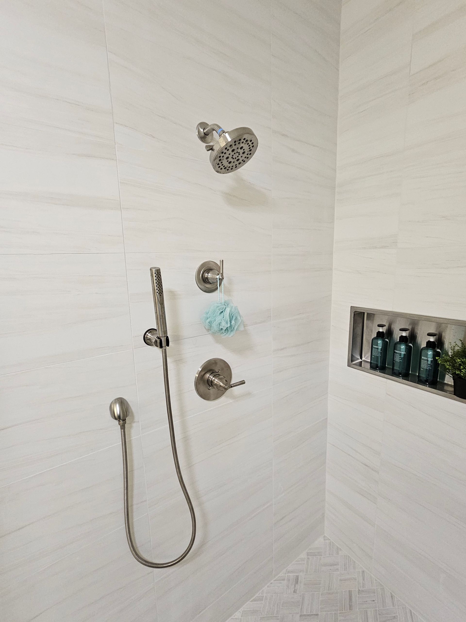 Brushed nickel shower fixtures against white tiled walls. A built-in shelf holds toiletries.