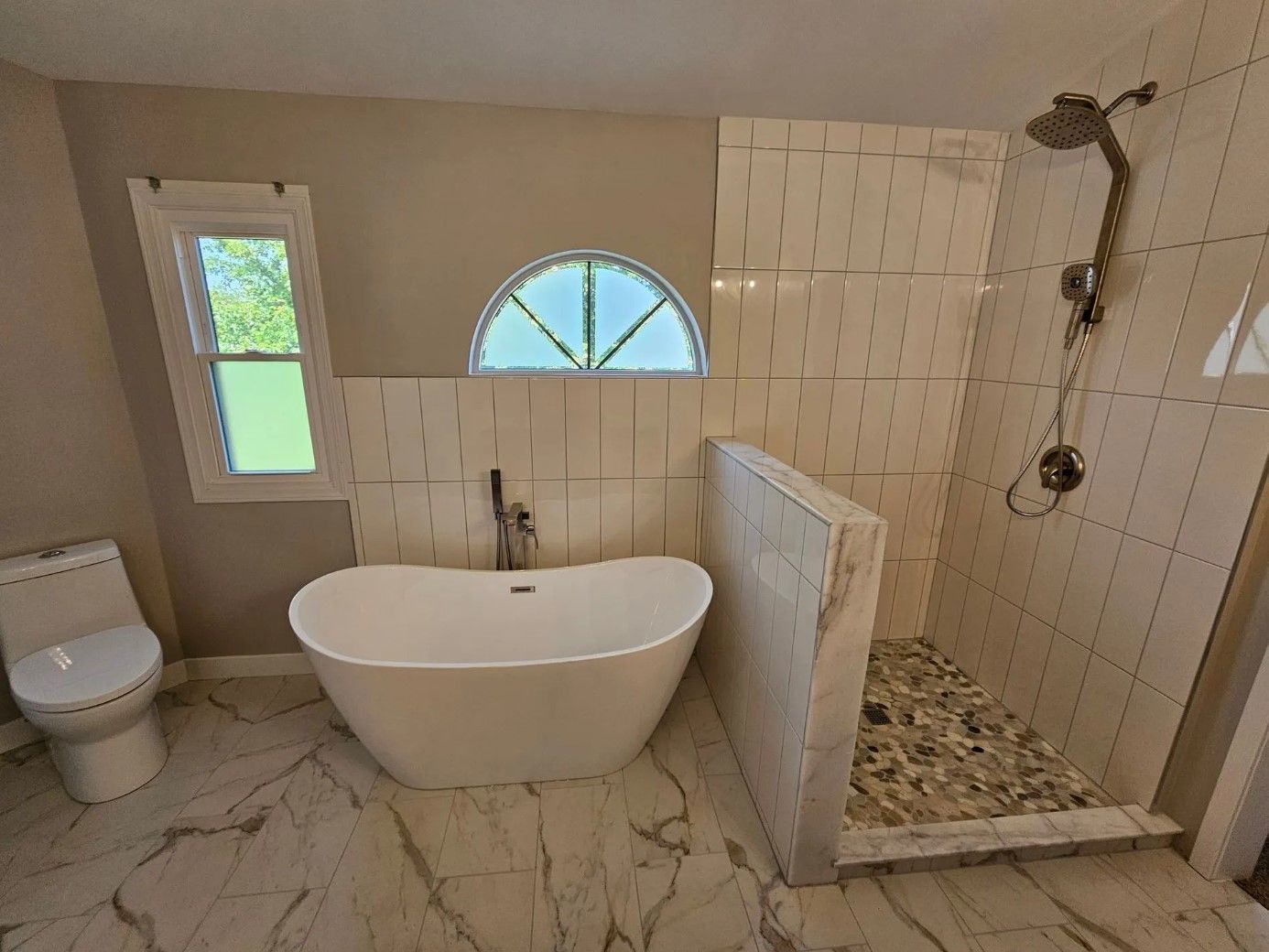 Bathroom with a freestanding tub, shower with pebble floor, toilet, and arched window.