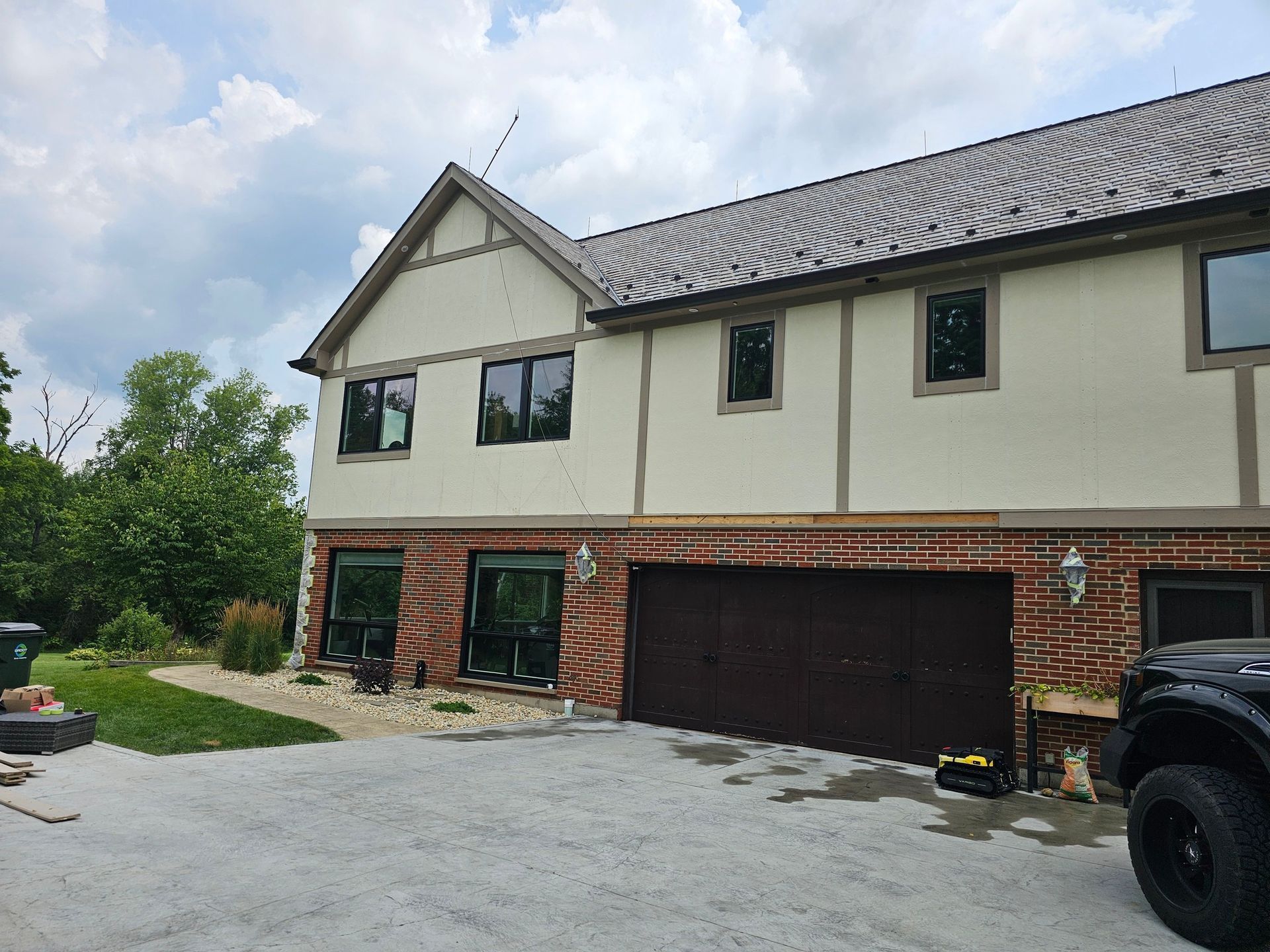 Two-story house with red brick and beige walls, brown garage door, and black-framed windows, surrounded by gravel.