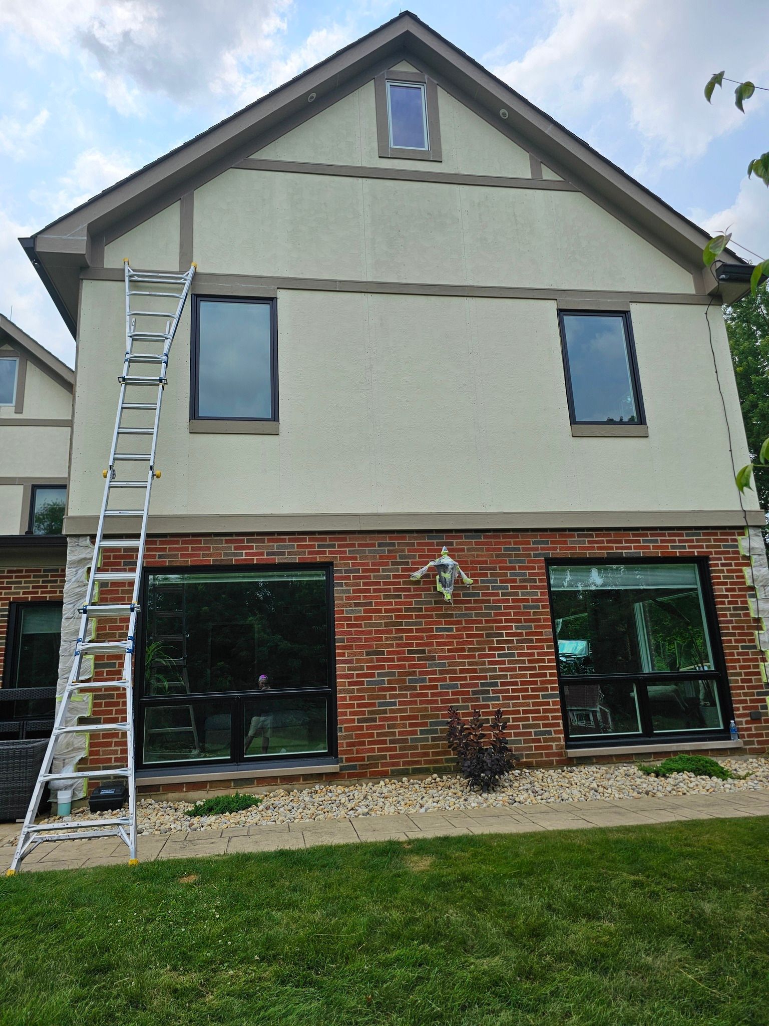Side of a two-story house with a ladder propped against it. Brick base, beige upper, black-framed windows.