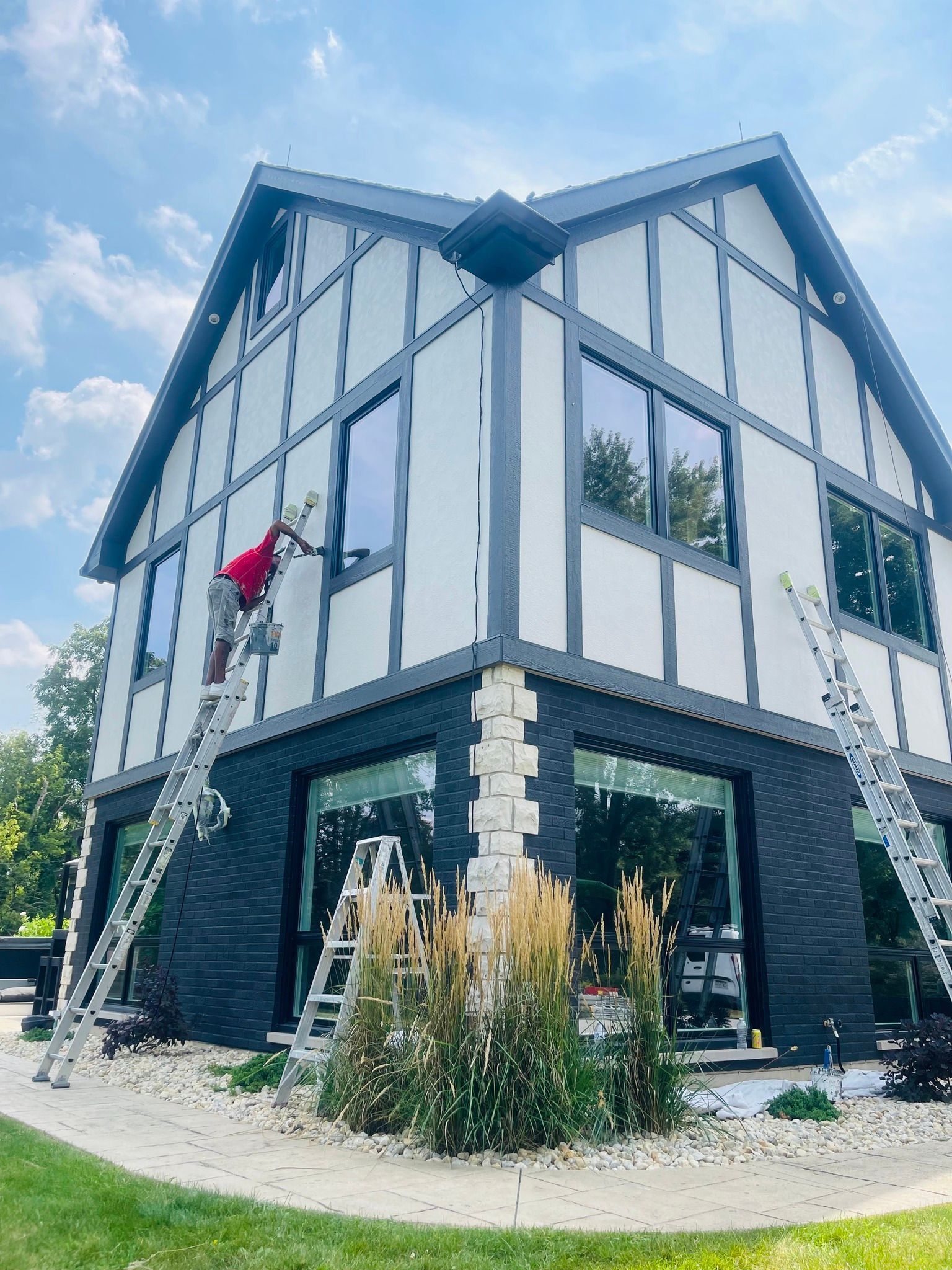 A person painting a two-story house with white siding, black trim, and large windows, from a ladder.