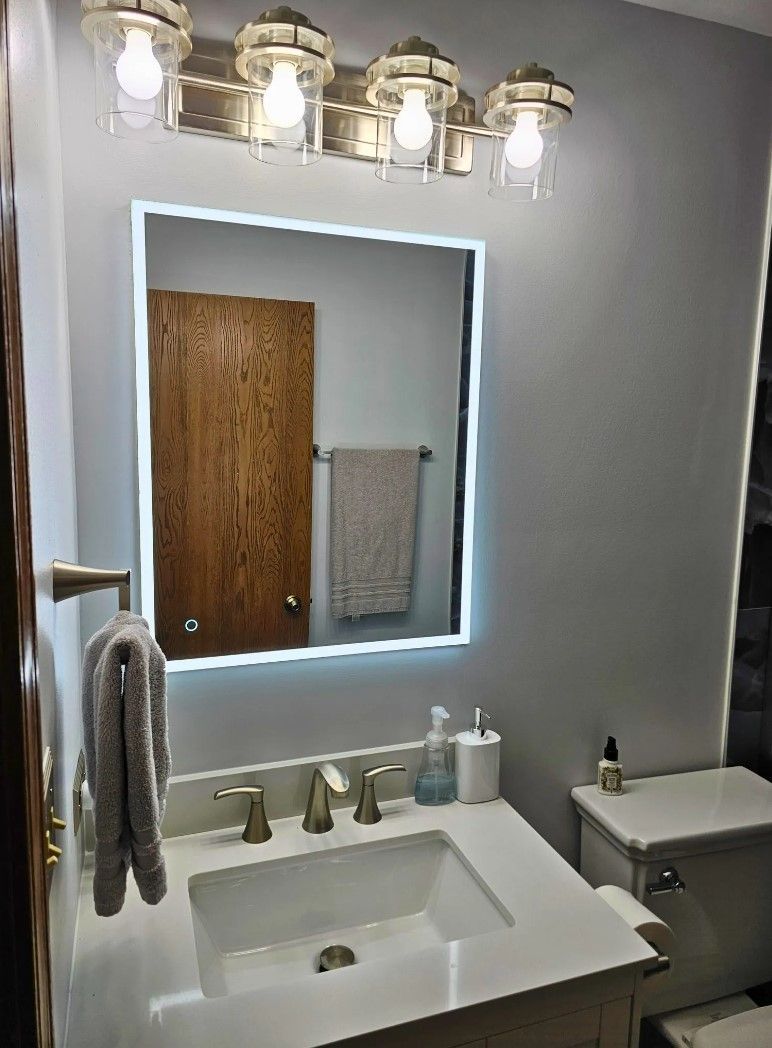 Bathroom with lighted mirror over a white sink and toilet; gray walls, gold fixtures, and wooden door.