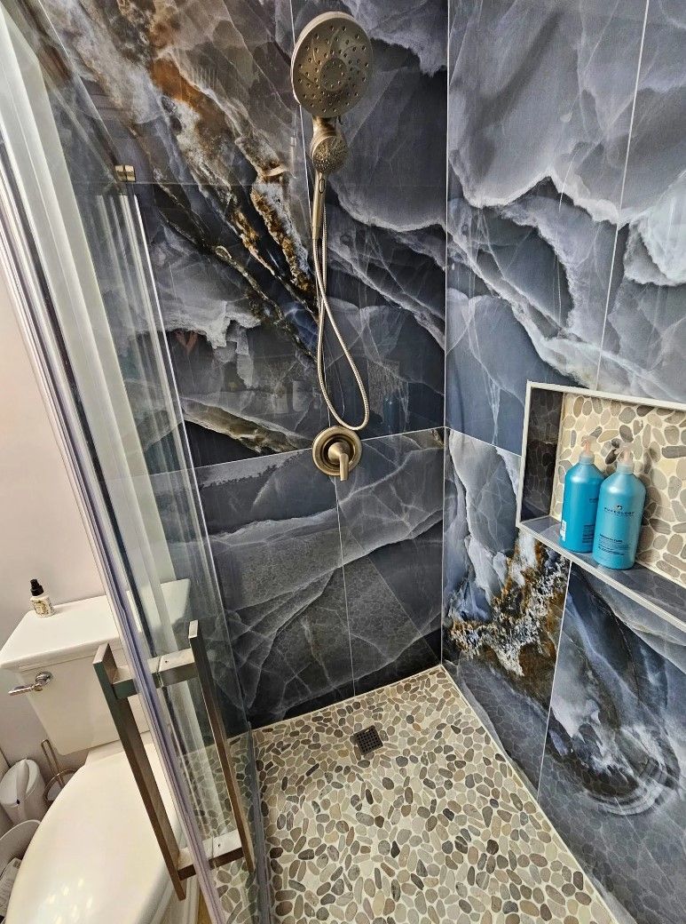 Shower with dark blue marble-like tiles, pebble floor, glass door, and built-in shelf with shampoo.