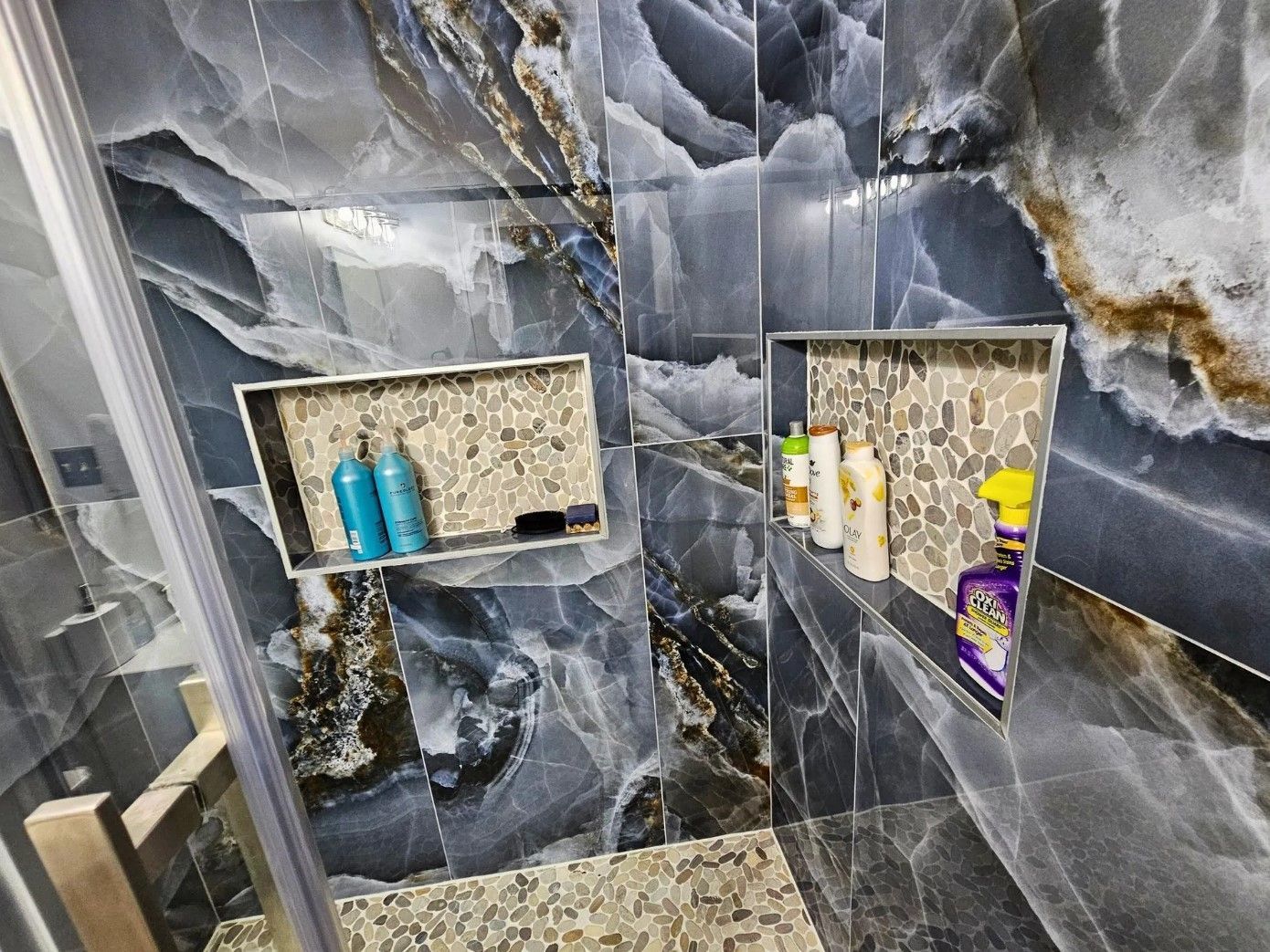 Shower with dark marbled walls, two built-in niches with toiletries, and a glass door.