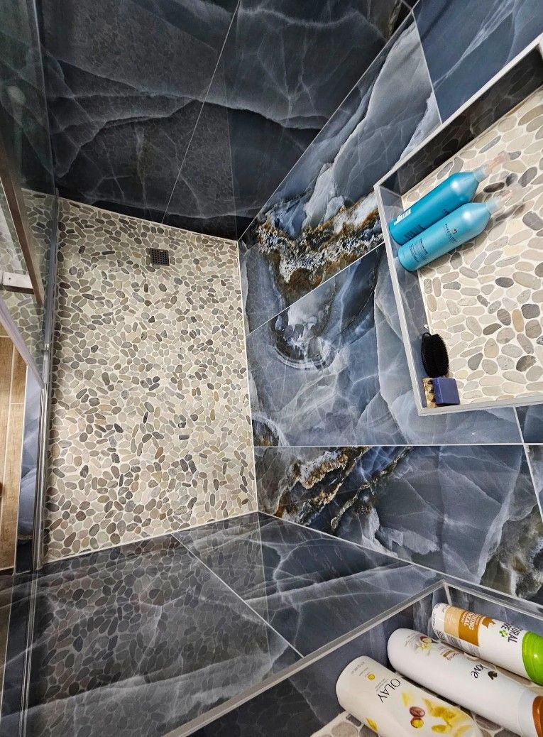 Shower with blue marble tile, pebble floor, and toiletries on a built-in shelf.