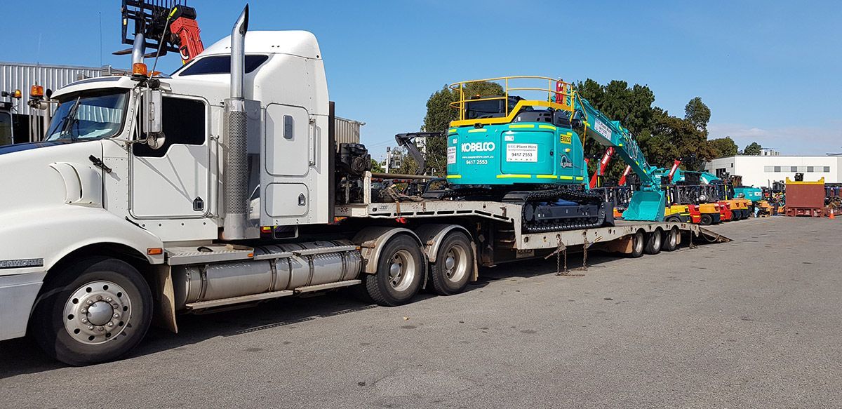 A white semi truck is carrying a blue excavator on a flatbed trailer.