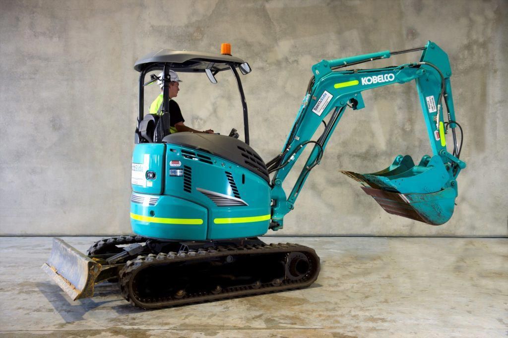 A man is driving a blue excavator on a concrete floor.