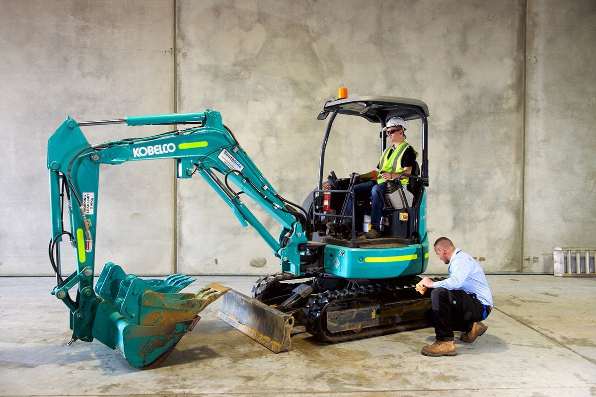 A man is kneeling next to a small excavator in a warehouse.