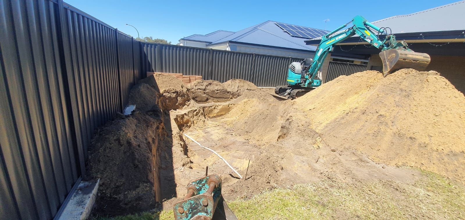 A green excavator is digging a hole in a backyard next to a pile of dirt.