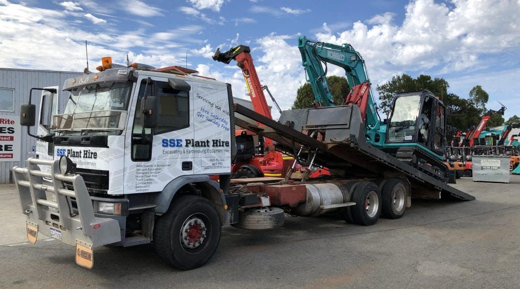 A tow truck is carrying a blue excavator on a flatbed trailer.