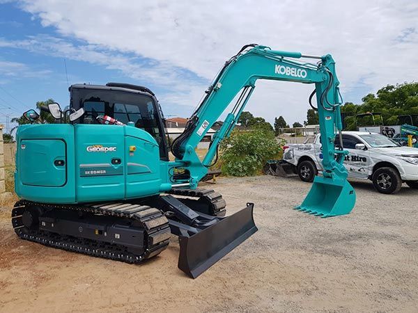A blue excavator is parked in a dirt lot next to a white truck.