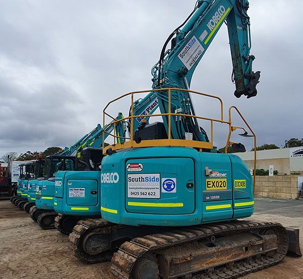 A row of blue excavators are parked in a lot.